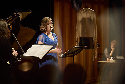 Studierende performen live auf der Bühne, im Vordergrund singt eine Sängerin in einem blauen Kleid