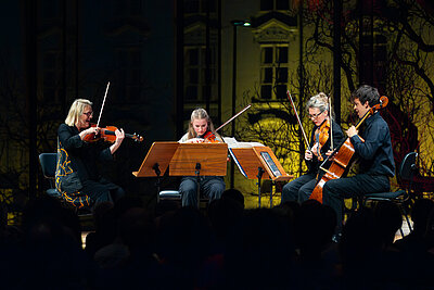 Ein Streicherensemble spielt live im Scheinwerferlicht auf einer Konzertbühne. Durch das große Fenster hinter der Bühne sieht man einen Teil der Hofburg.