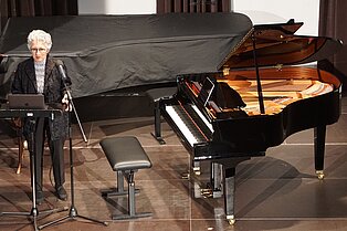 Prof.in Dr.in Carol Leone steht am Rednerpult auf der Bühne, neben dem Klavier mit verkleinerter Klaviatur. Ein Konzertflügel steht im Hintergrund.