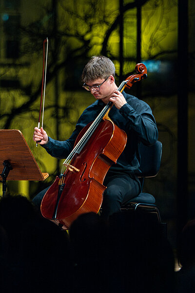 Ein Studierender spielt live Cello im Scheinwerferlicht auf einer Konzertbühne. Durch das große Fenster hinter der Bühne sieht man einen Teil der Hofburg.