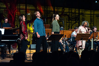 Jazzorchester mit Sänger*innen spielen live auf einer beleuchteten Konzertbühne