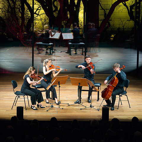 Ein Streicherensemble spielt live im Scheinwerferlicht auf einer Konzertbühne. Durch das große Fenster hinter der Bühne sieht man einen Teil der Hofburg.