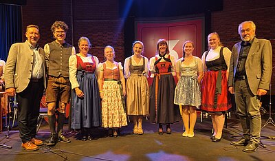 Studierende des Lehrgangs Alpenländische Volksmusikakademie, Lehrgangsleiter Peter Margreiter und Direktor Max Bauer stehen auf einer Bühne